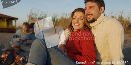 Image of Loving Young Couple Sitting On The Beach beside Campfire drinkin