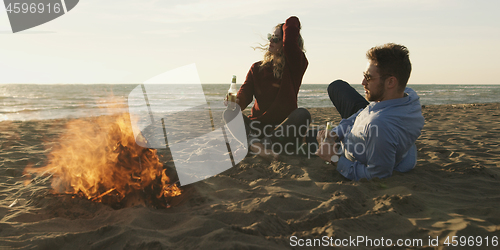 Image of Loving Young Couple Sitting On The Beach beside Campfire drinkin