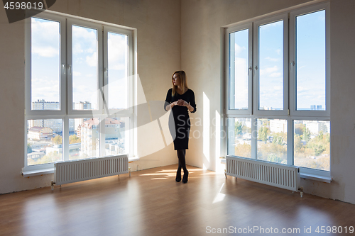 Image of A girl in a black dress stands between two large windows in a spacious empty apartment