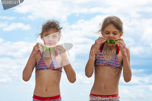 Image of Two girls eat watermelon on the beach against the cloudy sky