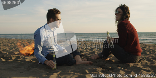 Image of Loving Young Couple Sitting On The Beach beside Campfire drinkin