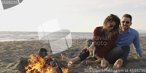 Image of Loving Young Couple Sitting On The Beach beside Campfire drinkin