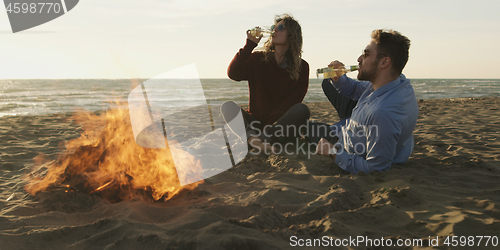 Image of Loving Young Couple Sitting On The Beach beside Campfire drinkin