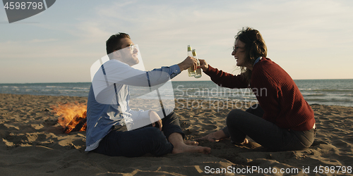 Image of Loving Young Couple Sitting On The Beach beside Campfire drinkin