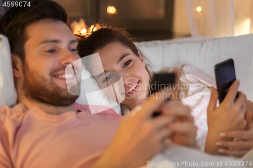 Image of happy couple using smartphones in bed at night