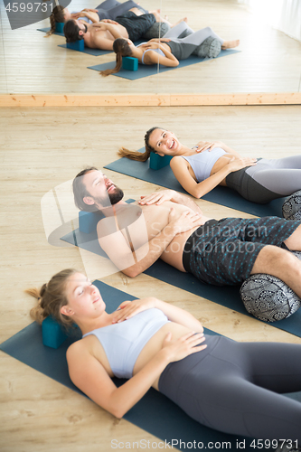 Image of Restorative yoga with a bolster. Group of young sporty attractive people in yoga studio, lying on bolster cushion, stretching and relaxing during restorative yoga. Healthy active lifestyle