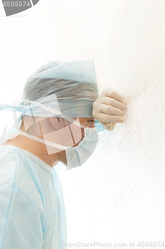 Image of Tired surgeon in sterile gown and mask leaned his head against the wall