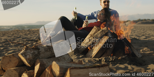 Image of Loving Young Couple Sitting On The Beach beside Campfire drinkin