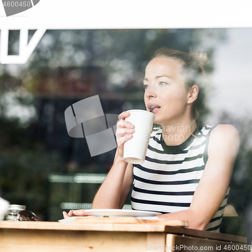 Image of Young caucasian woman sitting alone in coffee shop thoughtfully leaning on her hand, looking trough the window