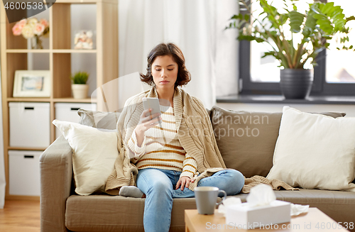 Image of sad sick woman in blanket using smartphone at home