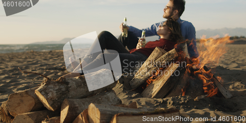 Image of Loving Young Couple Sitting On The Beach beside Campfire drinkin