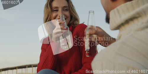 Image of Loving Young Couple Sitting On The Beach beside Campfire drinkin