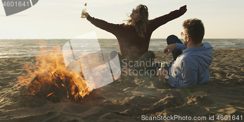 Image of Loving Young Couple Sitting On The Beach beside Campfire drinkin