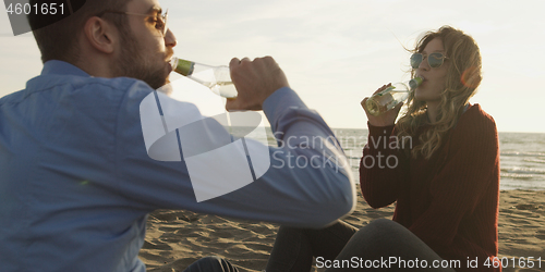 Image of Loving Young Couple Sitting On The Beach beside Campfire drinkin