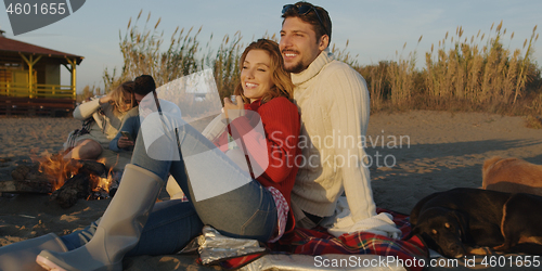 Image of Loving Young Couple Sitting On The Beach beside Campfire drinkin