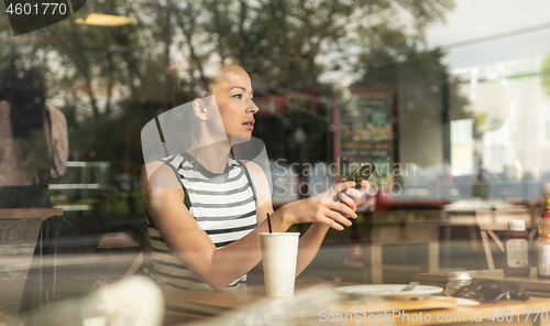 Image of Thoughtful caucasian woman holding mobile phone while looking through the coffee shop window during coffee break.