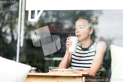 Image of Young caucasian woman sitting alone in coffee shop thoughtfully leaning on her hand, looking trough the window