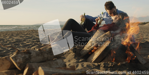 Image of Loving Young Couple Sitting On The Beach beside Campfire drinkin