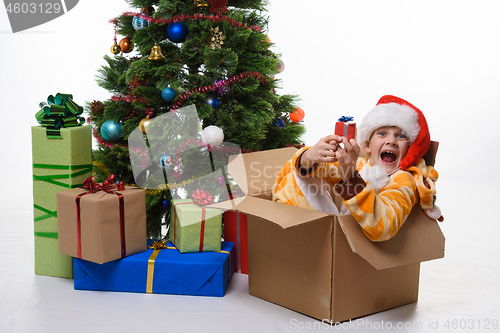 Image of The girl sits in a box of Christmas trees near the Christmas tree and cheerfully looks into the frame