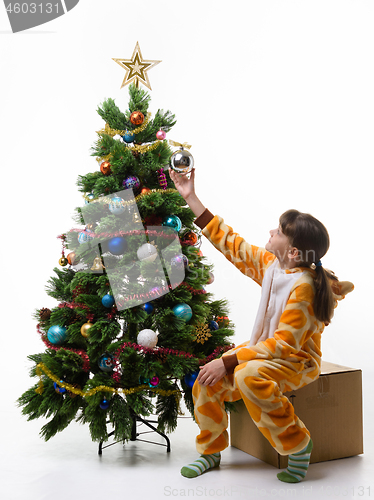 Image of The girl sits on a box in front of the Christmas tree and hangs a Christmas tree ball