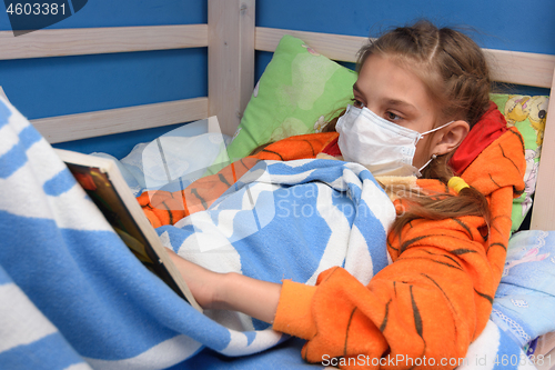 Image of Sick girl in medical mask lies in bed and reads a book