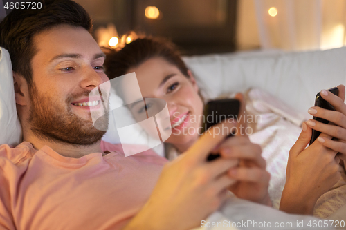 Image of happy couple using smartphones in bed at night