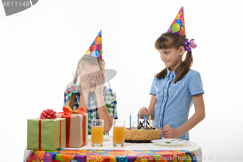 Image of While one girl inserts candles in a birthday cake, another girl covers her face with her palms