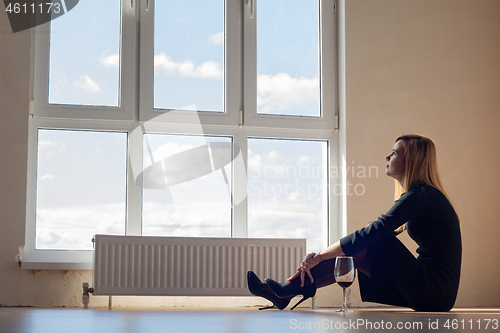 Image of Sad girl sitting by the window in an empty apartment with a glas