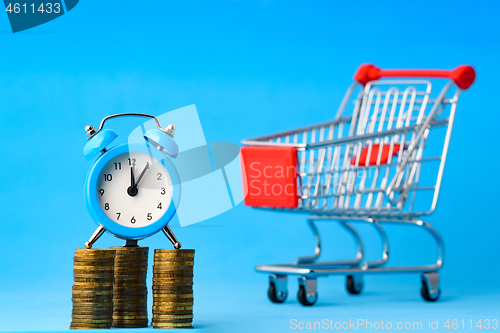 Image of A clock stands on a stack of coins, in the background a grocery cart