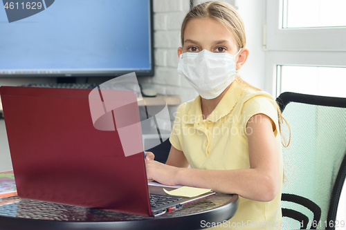 Image of A girl in a medical mask in a home setting learns remotely and looked into the frame