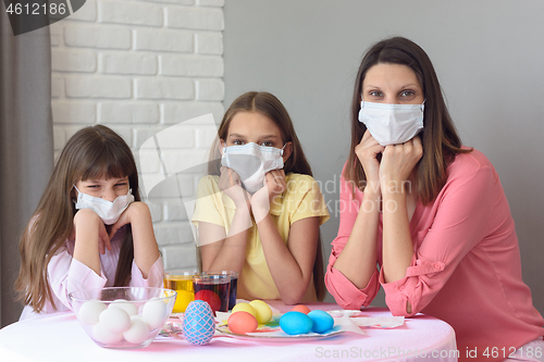 Image of Mom and two daughters at the table in medical masks paint Easter eggs for the holiday