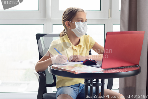 Image of A girl in a medical mask on quarantine in isolation isolates herself remotely from her lessons and distracted, looked out the window