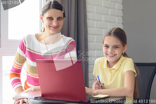 Image of Happy girl and mom with a smile look at the frame, sitting at the table and doing homework