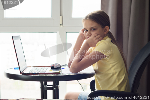 Image of An upset girl sits at a table at home, does homework and looks with displeasure