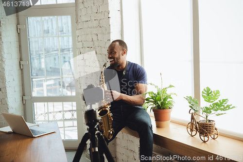 Image of African-american musician playing saxophone during online concert at home isolated and quarantined, laughting