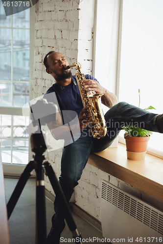 Image of African-american musician playing saxophone during online concert at home isolated and quarantined, impressive improvising