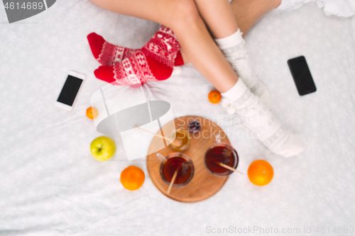 Image of Soft photo of woman and man on the bed with phone and fruits, top view point. Female and male legs in warm woolen socks