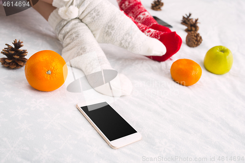 Image of Soft photo of woman and man on the bed with phone and fruits, top view point. Female and male legs in warm woolen socks