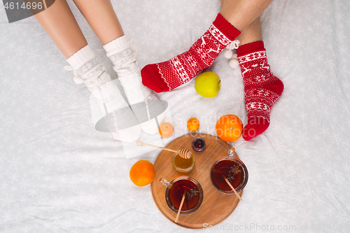 Image of Soft photo of woman and man on the bed with tea and fruits, top view point. Female and male legs in warm woolen socks