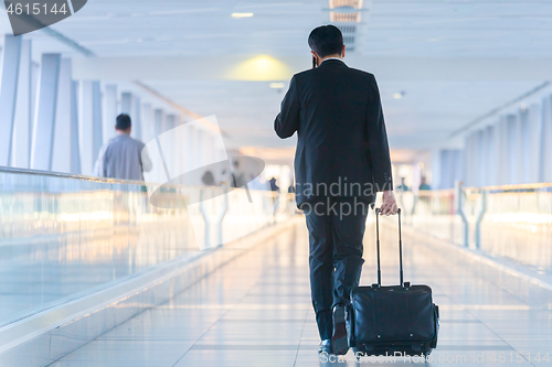 Image of Businessman walking and wheeling a trolley suitcase at the lobby, talking on a mobile phone. Business travel concept.