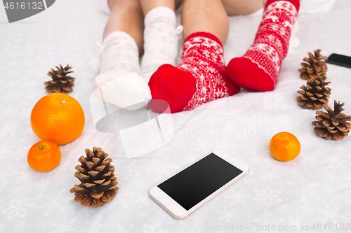 Image of Soft photo of woman and man on the bed with phone and fruits, top view point. Female and male legs in warm woolen socks