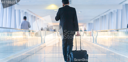 Image of Businessman walking and wheeling a trolley suitcase at the lobby, talking on a mobile phone. Business travel concept.