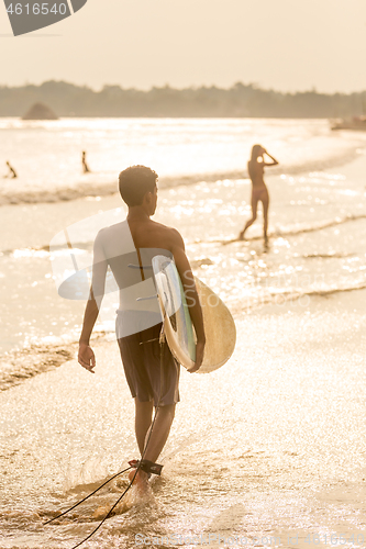 Image of Man walking at tropical beach of Midigama, Sri Lankha at sunsen with surf board in his hands.