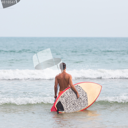 Image of Rear view of unrecognizable male surfer at tropical beach with sup surf board and paddle in his hands