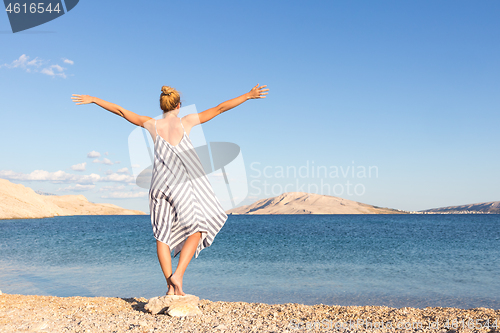 Image of Happy carefree woman rising arms, wearing beautiful striped summer dress enjoying late afternoon on white pabbled beach on Pag island, Croatia