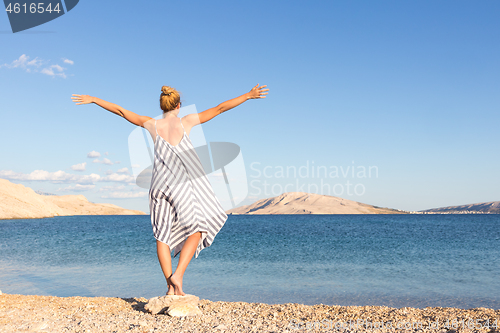 Image of Happy carefree woman rising arms, wearing beautiful striped summer dress enjoying late afternoon on white pabbled beach on Pag island, Croatia