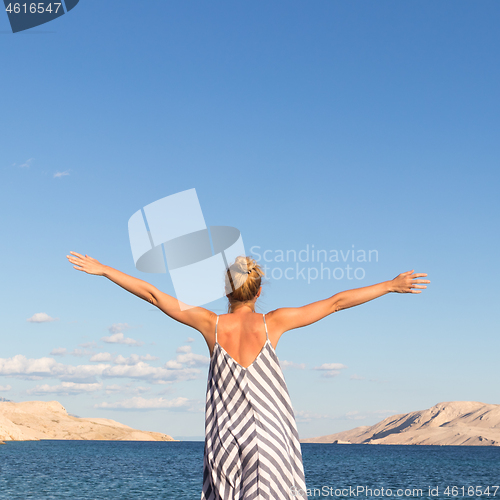 Image of Happy carefree woman rising arms, wearing beautiful striped summer dress enjoying late afternoon on white pabbled beach on Pag island, Croatia