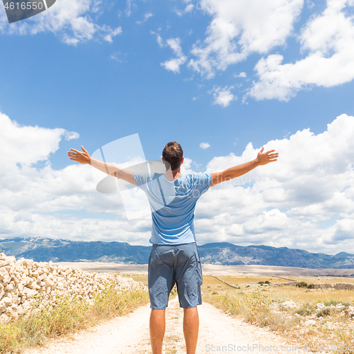 Image of Rear view of casual sporty man standing on a dirt country road rising hands up to the clouds on a blue summer sky. Freedom and travel adventure concept.