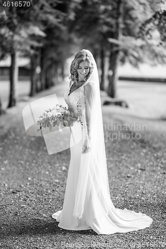 Image of Full length portrait of beautiful sensual young blond bride in long white wedding dress and veil, holding bouquet outdoors in natural background.