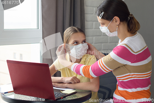 Image of Mom puts on a girl a medical face mask while sitting in front of a computer at home